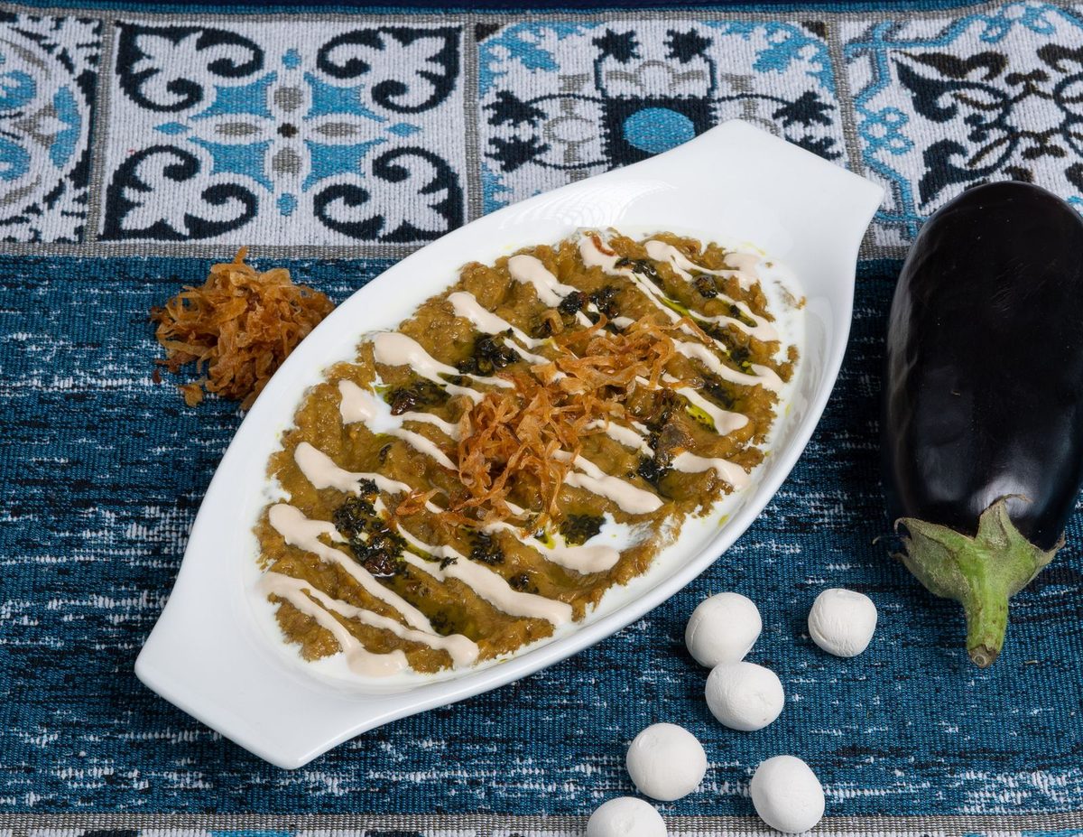 A plated cooked eggplant dish with a creamy topping, herbs, and nuts.