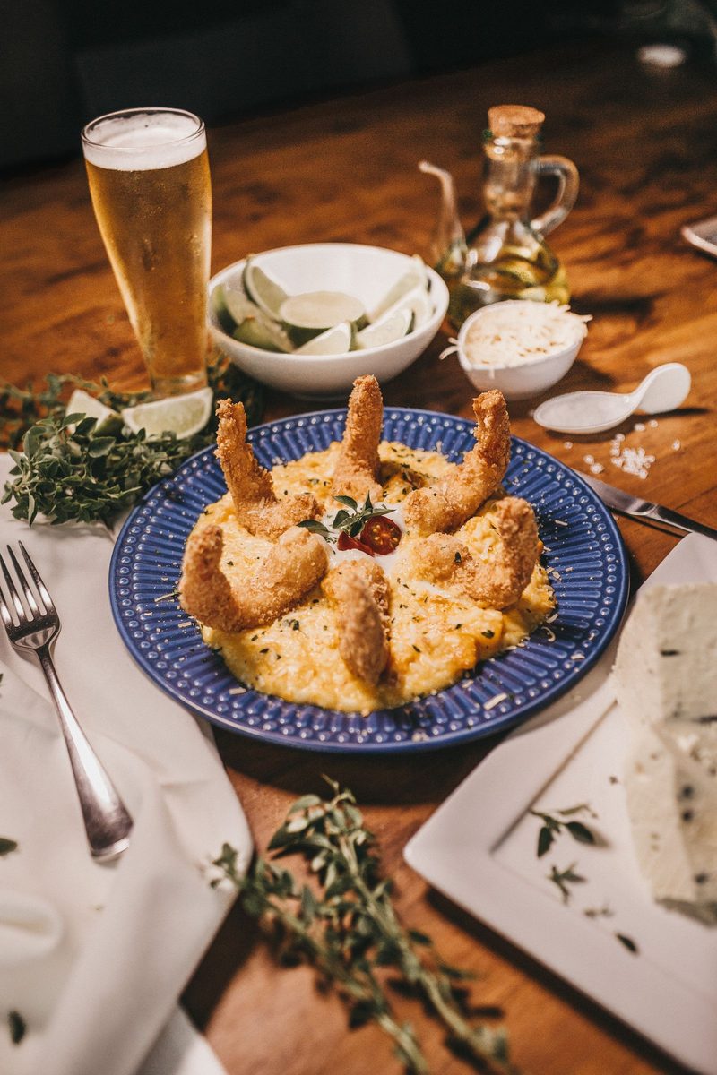 A plate of creamy risotto topped with fried shrimp beside a beer on a rustic wooden table