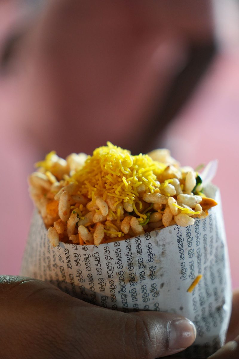 Close-up of bhel puri with puffed rice, sev, peanuts, and vegetables served in a folded newspaper cone held by hand.