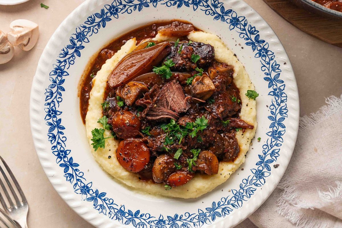 Finished French boeuf bourguignon: tender beef and vegetables in a rich brown wine sauce, garnished with fresh herbs and served over creamy purée.