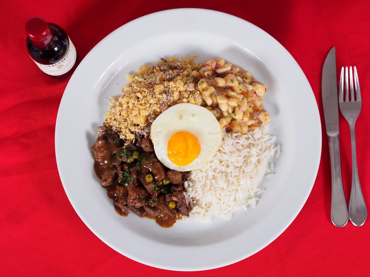Brazilian feijoada with black beans and sliced sausage served beside white rice and a fried egg on a white plate against a red background.
