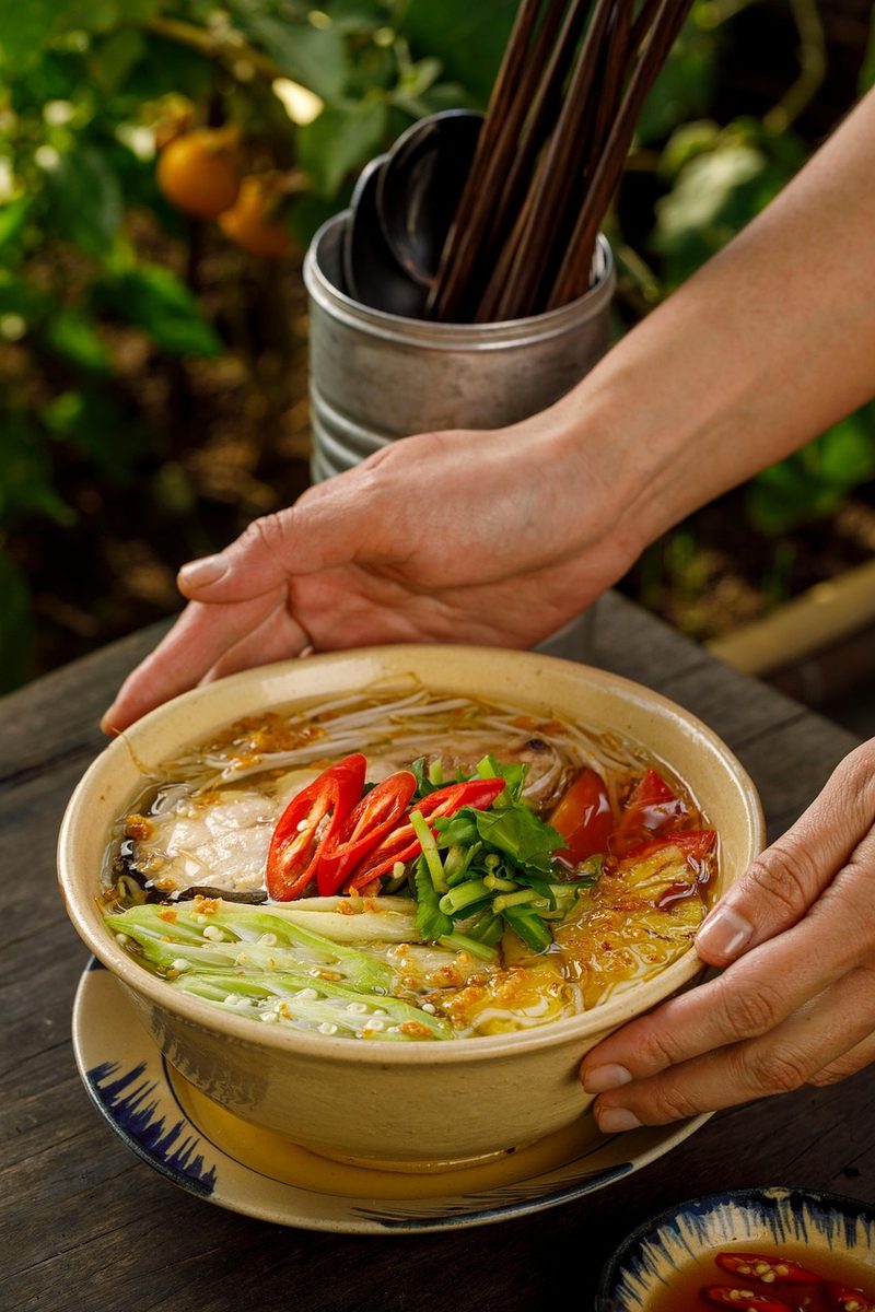 Finished bowl of Canh Chua Vietnamese sweet-and-sour soup with red chilies, scallions, herbs, vegetables, and seafood in a clear broth, presented outdoors with chopsticks nearby.