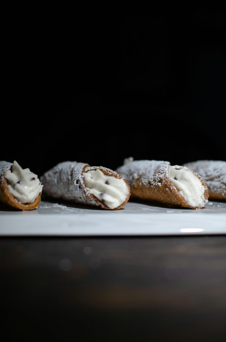 Several powdered-sugar-dusted Sicilian cannoli filled with white ricotta cream arranged on a white plate against a dark background.