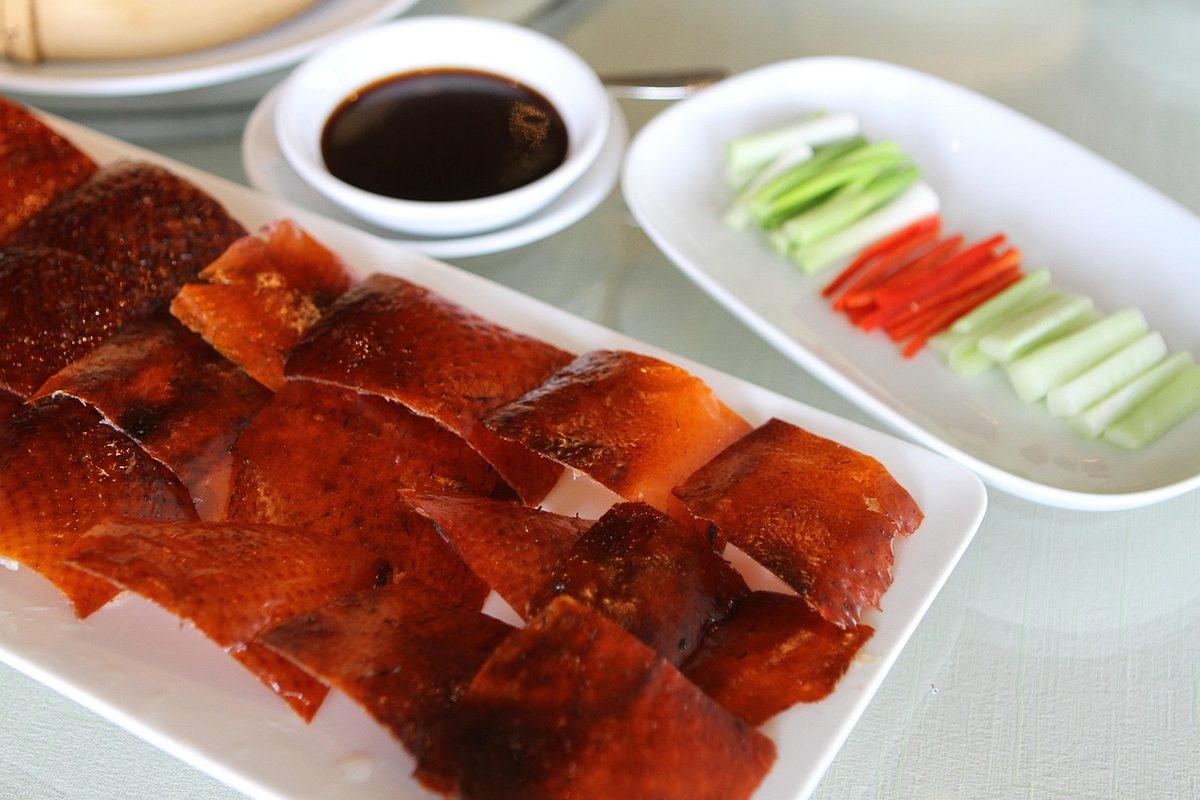 Sliced roast duck arranged on plates with cucumber and carrot sticks, scallions, pancakes, and dipping sauce.