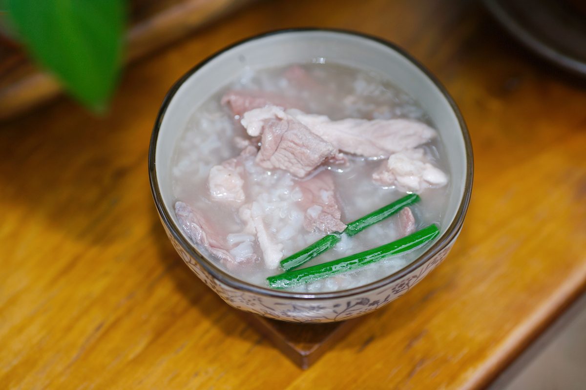 Bowl of Chinese rice porridge (congee) with pork pieces and green onion garnish on a wooden tray.