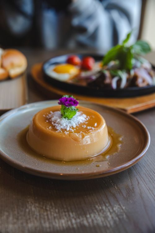 A single caramel flan, a smooth golden custard dessert topped with caramel sauce, powdered sugar, mint, and a small purple flower, served on a gray plate over a wooden table.