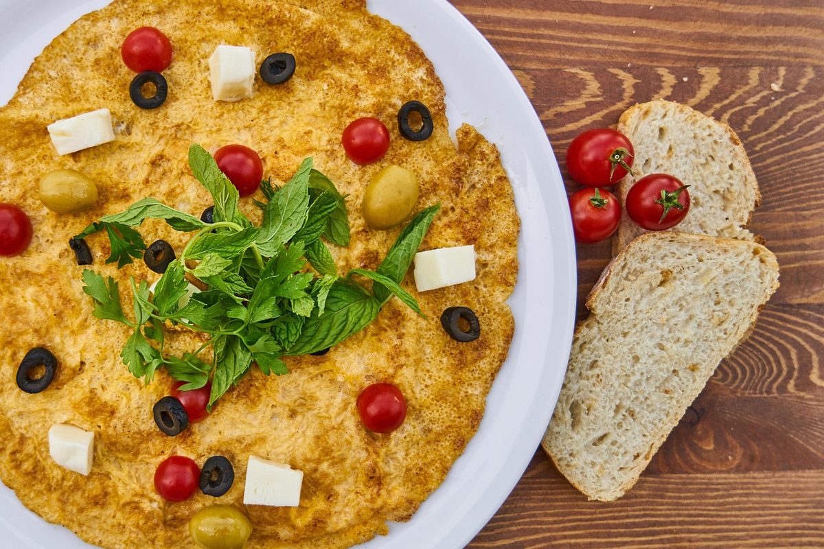 A large flat golden omelet on a white plate, topped with cherry tomatoes, black and green olives, cheese cubes, and fresh herbs, with sliced bread beside it on a wooden table.