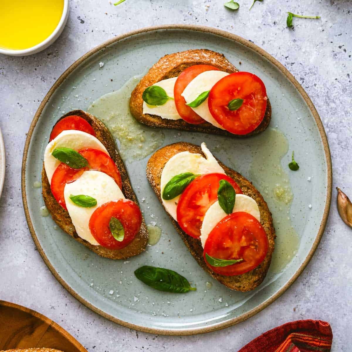 Three slices of toasted bread layered with fresh mozzarella and ripe tomato slices, garnished with basil and olive oil on a gray ceramic plate.