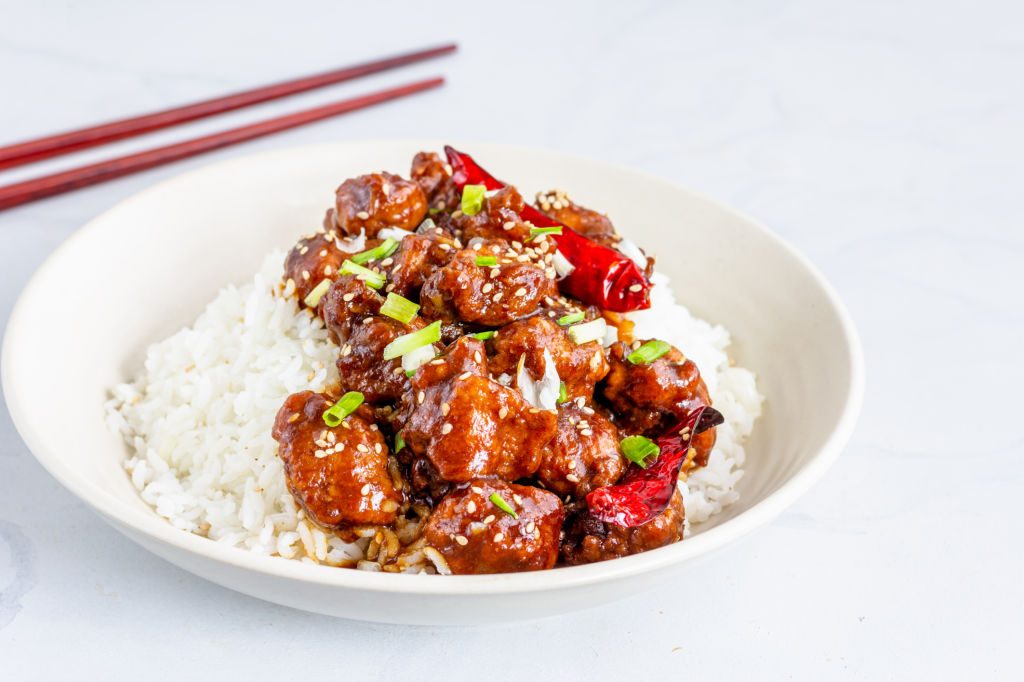 Glazed General Tso's chicken pieces garnished with sesame seeds, scallions, and red chilies, served over white rice in a bowl with chopsticks nearby.