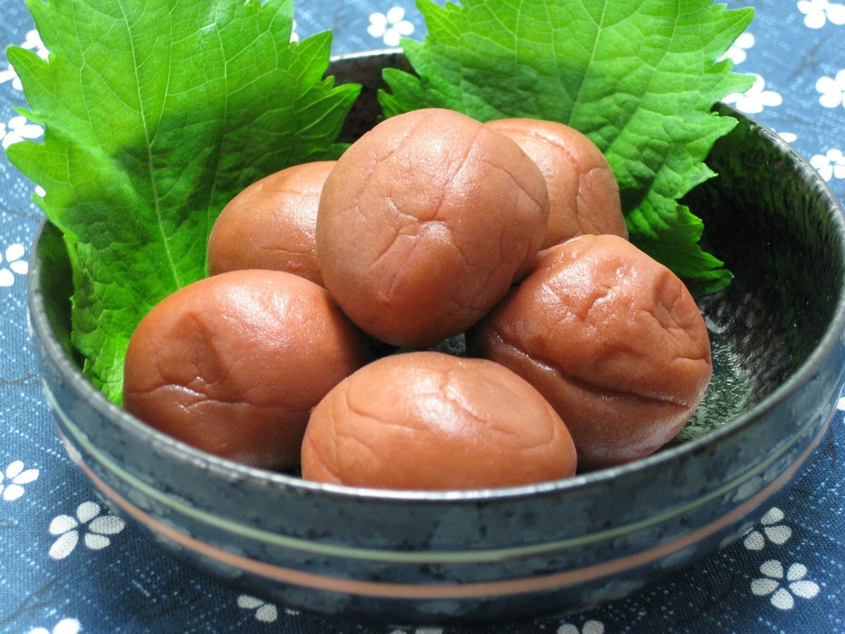 Several pinkish-brown umeboshi pickled plums arranged in a black bowl with green shiso leaves.