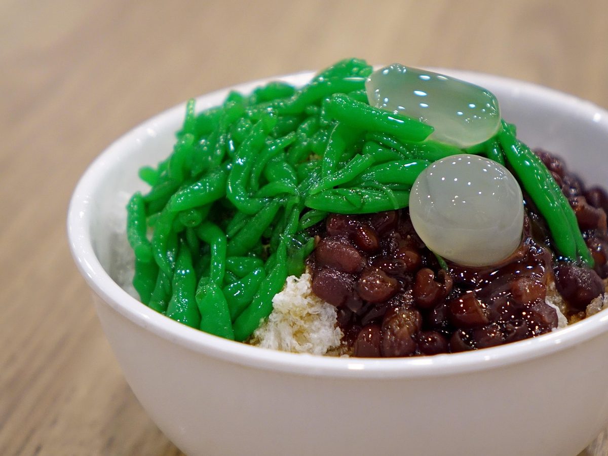 Bowl of green cendol noodles with coconut milk, ice, and red beans, photographed as a finished Southeast Asian dessert.