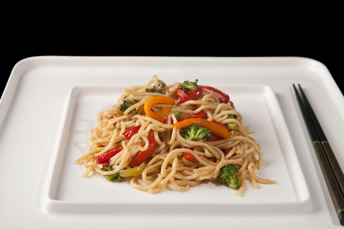 A plated serving of stir-fried Asian vegetable noodles with broccoli and red, orange, and green peppers on a white square plate, with chopsticks beside it.