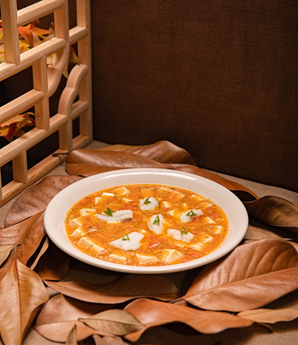 A white bowl of orange-red mapo tofu with tofu cubes, red sauce, and green onion garnish, surrounded by autumn leaves.