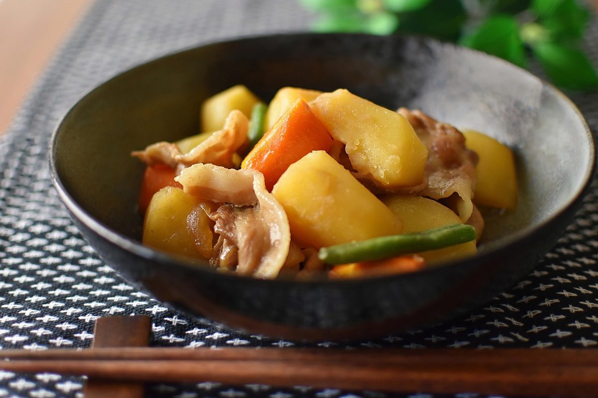 A bowl of Japanese nikujaga stew with tender meat, potatoes, carrots, and green beans in a savory broth, served with chopsticks on a dark patterned table.