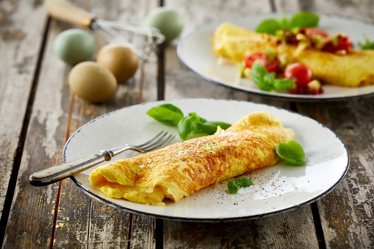 A golden rolled omelette served on a white plate with basil, pepper, and salt on a rustic wooden table.