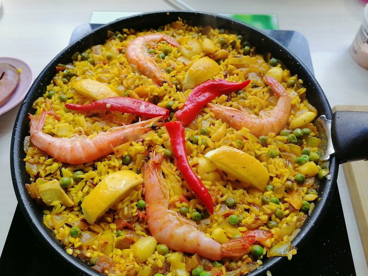 Finished Spanish seafood paella in a black pan, with saffron rice, whole shrimp, peas, lemon wedges, and red peppers.