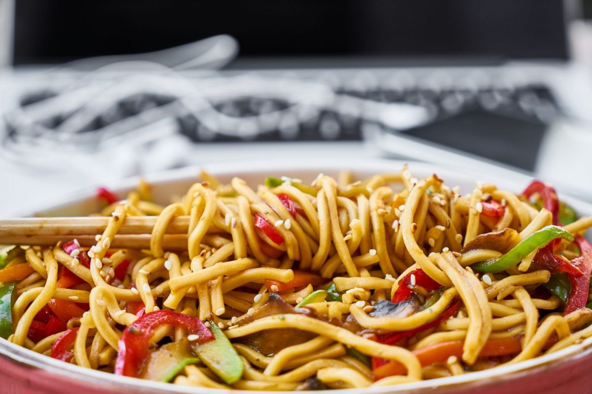 Colorful stir-fried noodles with vegetables and sesame seeds served in a white bowl.