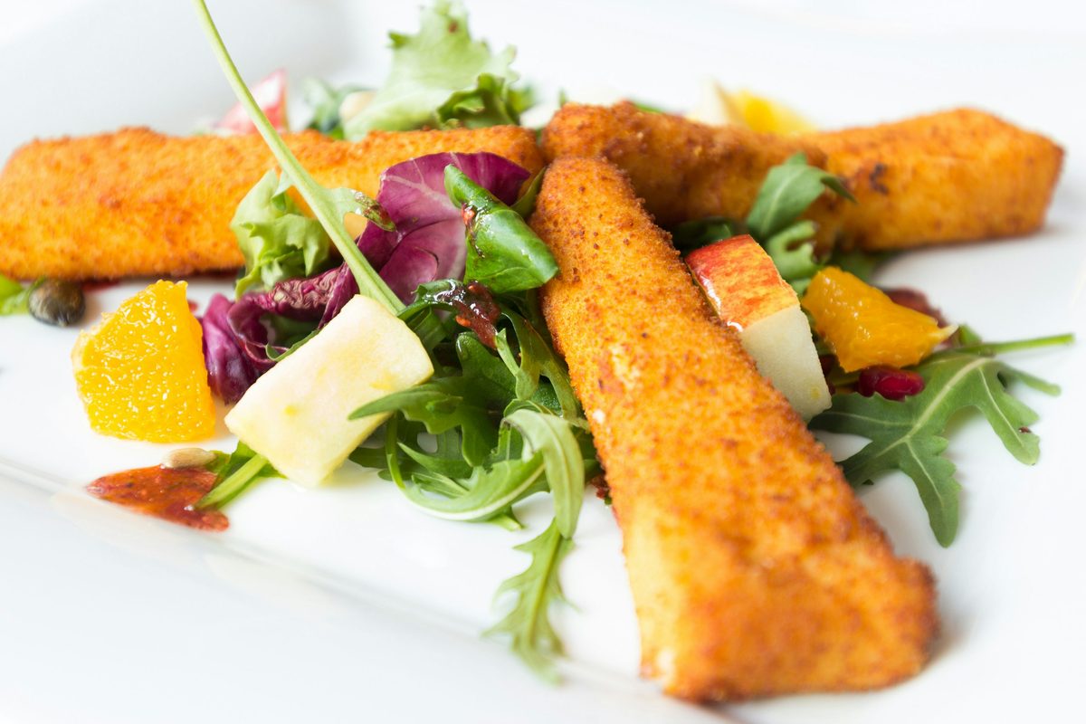 Golden fried saganaki cheese sticks arranged over fresh arugula and mixed greens with orange and apple pieces on a white plate.