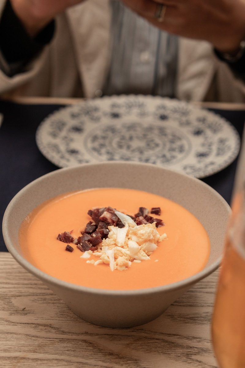 A bowl of creamy orange-red salmorejo topped with Iberian ham and chopped boiled egg, served in a ceramic bowl.