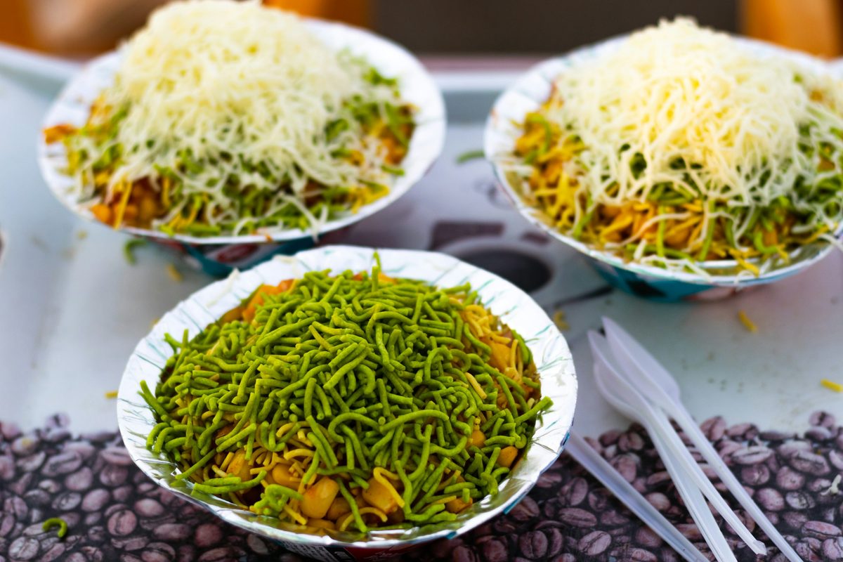 Three foil bowls of sev puri, an Indian chaat snack with potato filling, chutney, herbs, and sev, served as street food.