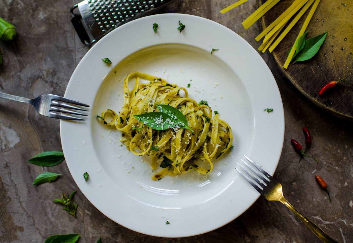 Top-down photograph of green pesto-coated flat pasta on a white plate, garnished with grated cheese and fresh basil, with a fork, grater, dry pasta, and red chilies arranged around it.