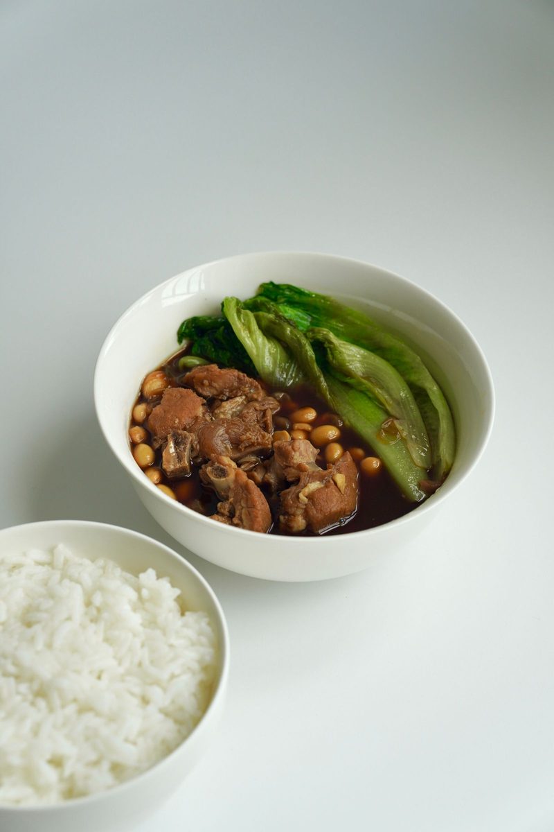 Braised pork pieces and tender mustard greens in dark broth, served with white rice in matching bowls.
