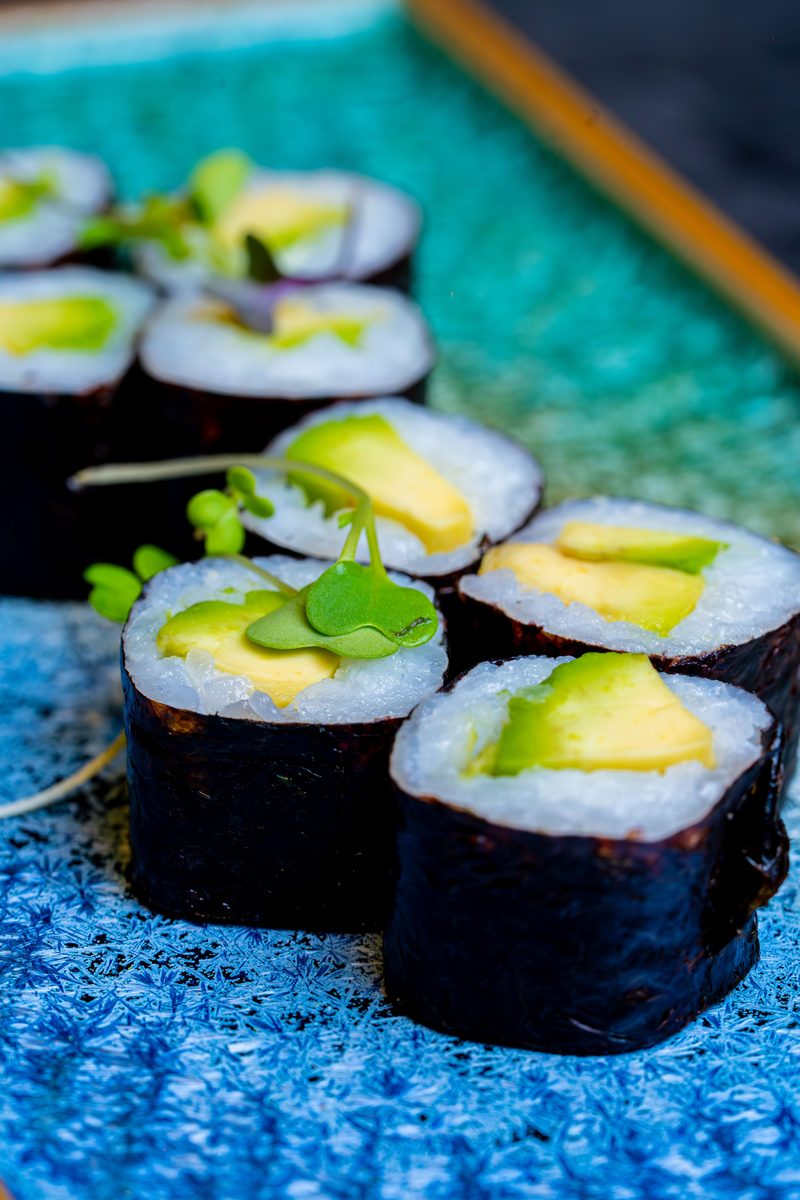 Sliced vegan maki rolls filled with avocado and garnished with microgreens, plated on a blue textured surface.