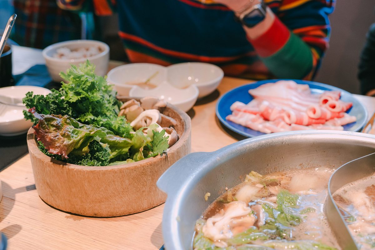 Japanese hot-pot meal with cooked greens, mushrooms, seafood, and broth in the foreground, with additional ingredients served at the table.