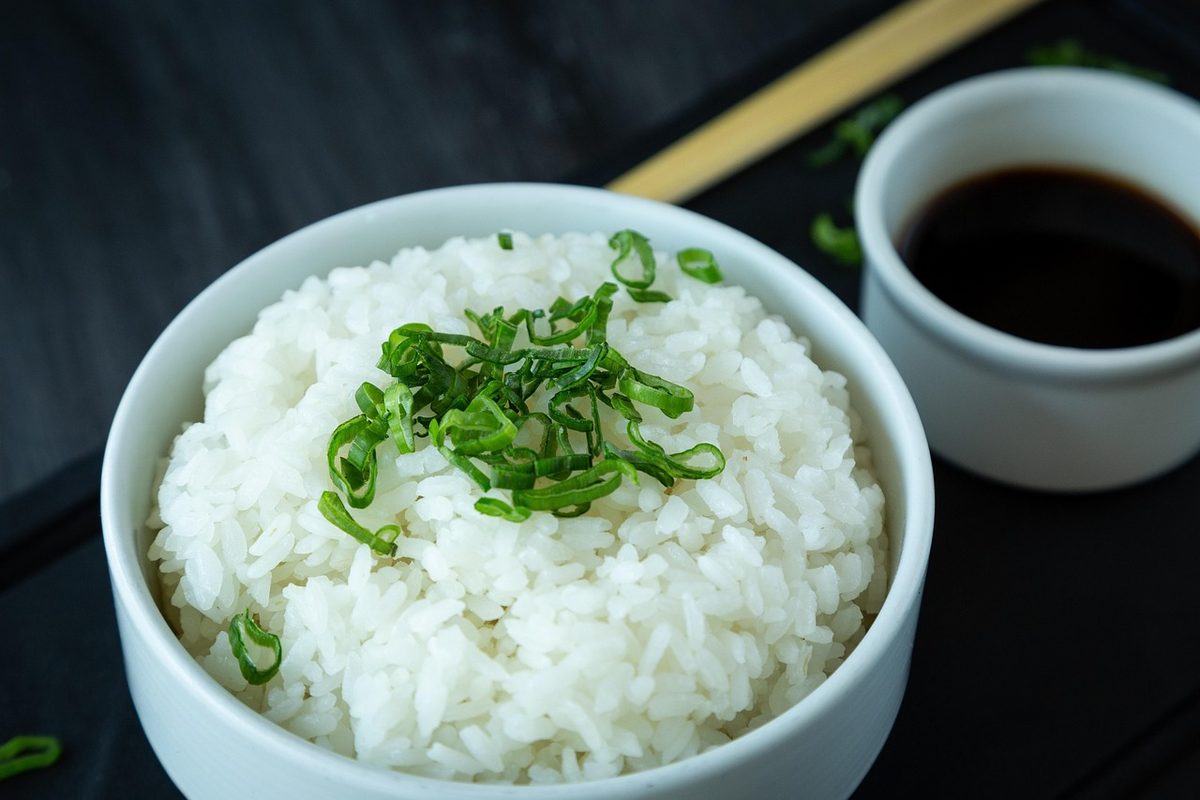 A white bowl of cooked Japanese rice topped with sliced green onions, with a small cup of soy sauce beside it.