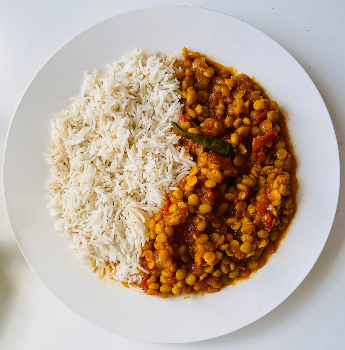 Top-down photograph of a finished golden-yellow lentil curry with tomato and a green chili beside fluffy white basmati rice on a white plate.