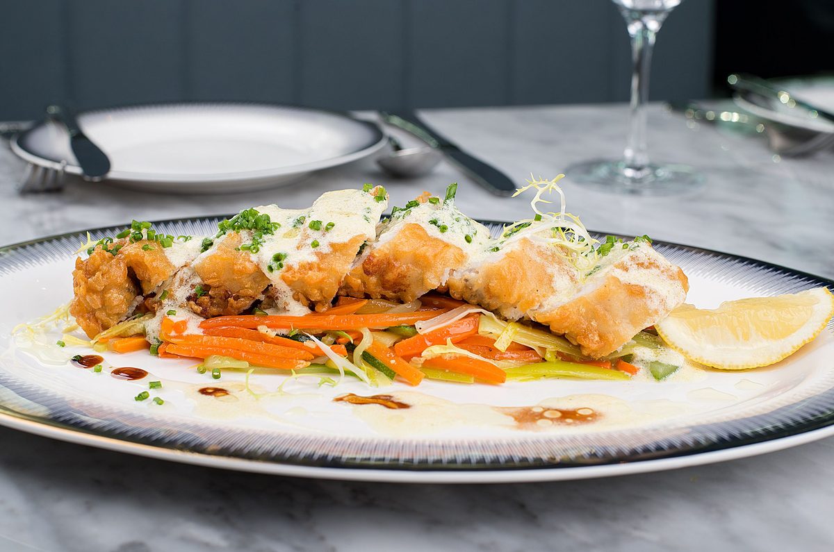 Crisp golden-battered fish pieces over sliced vegetables, finished with creamy herb sauce and lemon on a white plate.