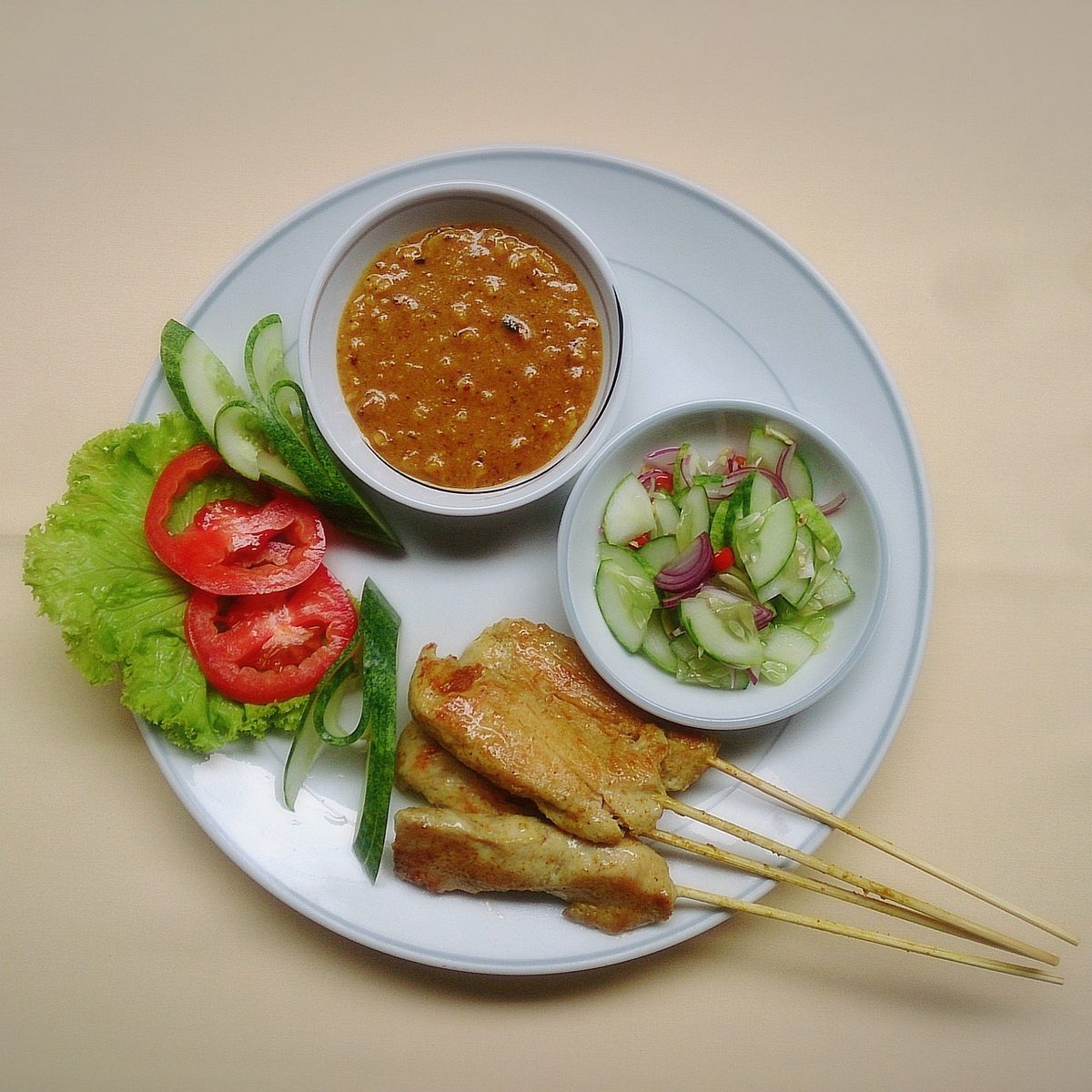 Thai chicken satay skewers served on a white plate with peanut dipping sauce, cucumber-onion relish, lettuce, and tomato slices.