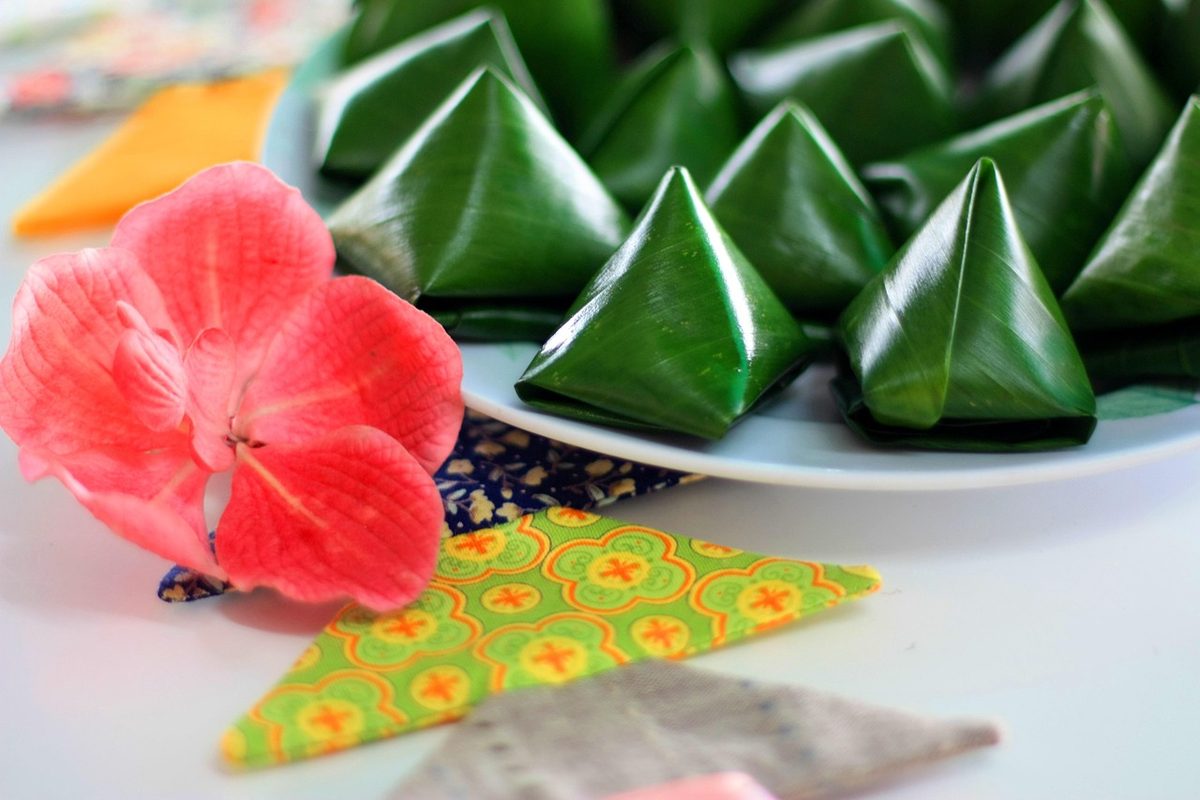 Finished Thai dessert of glossy green pandan-wrapped sweets arranged on a white plate with a pink tropical flower and patterned napkin.