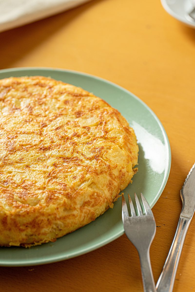 Whole golden Spanish tortilla de patatas (potato omelette) on a pale green plate with a fork, photographed against a yellow background.