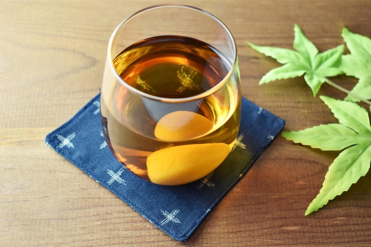 Glass of amber ume-shu plum wine on a blue coaster beside green plum leaves on a wooden table